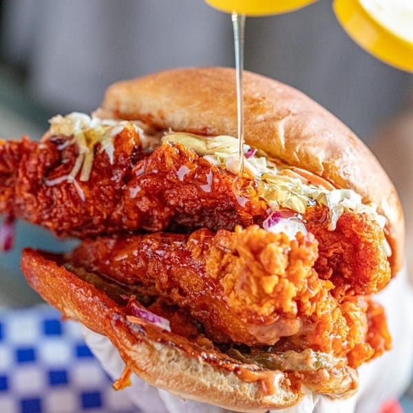 Honey being drizzled over a Legend Hot Chicken sandwich with crispy chicken, coleslaw, and sauce.